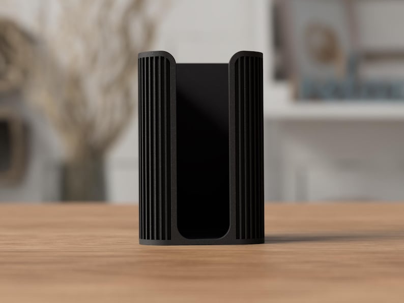 May include: A black, rectangular holder with vertical ribbed detailing on the sides. The holder is made of a matte material and sits on a light brown wooden surface. The background is blurred, suggesting an indoor setting.