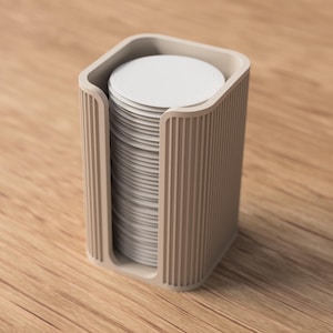May include: A beige, rectangular coaster holder with a stack of white coasters. The holder has a ribbed design on the sides and a curved top. The coasters are round and stacked neatly inside the holder. The holder is sitting on a wooden surface.