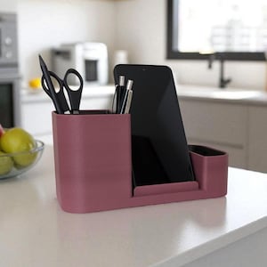 A dark pink desk organizer with a smartphone, pens, and scissors sits on a white kitchen counter with a blurred background.