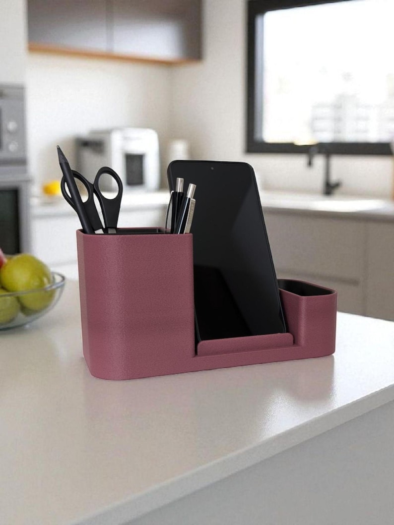 A dark pink desk organizer with a smartphone, pens, and scissors sits on a white kitchen counter with a blurred background.