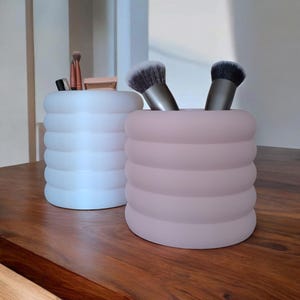 Könnte beinhalten: Zwei dekorative Behälter, einer weiß und einer rosa, stehen auf einer Holzoberfläche. Die Behälter haben ein geripptes Design und enthalten Make-up-Pinsel und andere Schönheitsprodukte. Die Behälter sind zylindrisch.