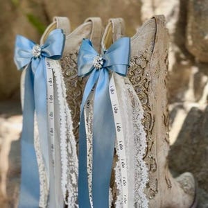Op de afbeelding: Drie beige cowboylaarzen met ingewikkelde borduursels en kanten details. Elke laars is versierd met een lichtblauwe strik en linten. Op de linten staat de tekst "I do". Een sprankelend juweel bevindt zich in het midden van elke strik.