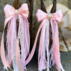 May include: Two brown leather cowboy boots adorned with decorative boot clips. Each clip features a large light pink satin bow centered with a faux pearl and rhinestone embellishment, with cascading ribbons of lace, gingham, and velvet.
