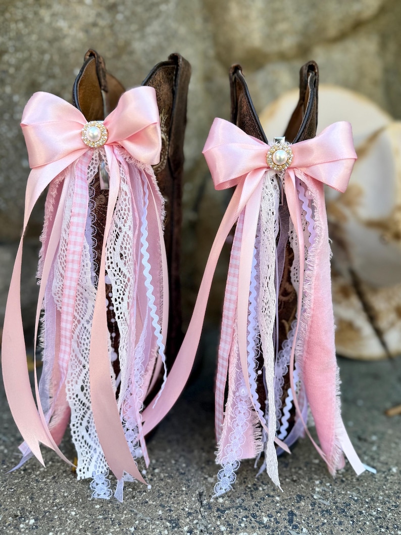 May include: Two brown leather cowboy boots adorned with decorative boot clips. Each clip features a large light pink satin bow centered with a faux pearl and rhinestone embellishment, with cascading ribbons of lace, gingham, and velvet.