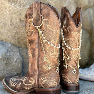May include: A pair of distressed brown leather cowboy boots featuring intricate gold-toned embroidery and decorative gold-tone chain accessories. The chains are adorned with small white faux pearls, tiny star charms, and a small heart-shaped pendant.