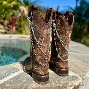 Puede incluir: Un par de botas de vaquero de cuero marrón con intrincados diseños bordados y detalles de cadena plateada. Las botas presentan un dije de cruz y están sobre un fondo exterior borroso.