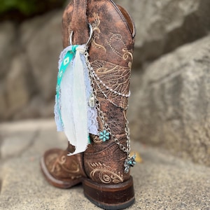 May include: Brown leather cowboy boot with decorative stitching and a silver chain with turquoise accents. The boot has a decorative fabric and lace charm attached to the top.