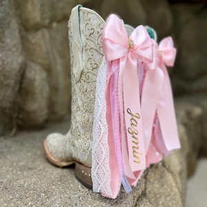 May include: A cream-colored cowboy boot featuring an intricate embossed pattern, adorned with layered pink satin bows, white lace trim, and a pink ribbon printed with the name Jazmin in gold script, accented with small rhinestones on the heel.
