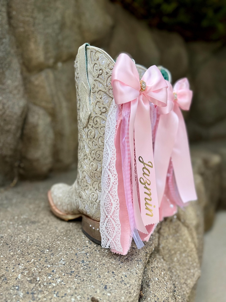 May include: A cream-colored cowboy boot featuring an intricate embossed pattern, adorned with layered pink satin bows, white lace trim, and a pink ribbon printed with the name Jazmin in gold script, accented with small rhinestones on the heel.