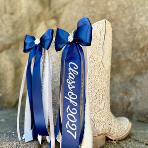 May include: A cream-colored cowboy boot with intricate floral patterns is adorned with navy blue ribbon bows and streamers. One ribbon is embroidered with "Class of 2027" in white. The bows have a rhinestone embellishment.