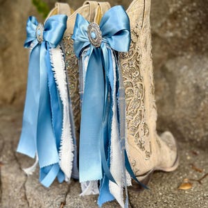 Op de afbeelding: Een paar beige cowboylaarzen met ingewikkelde borduursels en versieringen. Elke laars is versierd met een lichtblauwe satijnen strik, een zilverkleurig decoratief stuk en stromende linten. De laarzen staan tegen een natuurlijke achtergrond.