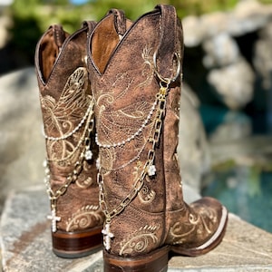 Puede incluir: Un par de botas de vaquero de cuero marrón con intrincados diseños florales bordados. Las botas presentan cadenas doradas decorativas con dijes de cruz y flores. Las botas tienen punta cuadrada y tacón apilado. Aproximadamente 30 cm de altura.