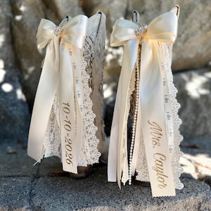 May include: A pair of cream-colored boots adorned with white lace trim and large satin bows. Each boot features hanging ribbons printed with the repeated text 'i do', one displaying the date '10-10-2026' and the other 'Mrs. Caylor' in gold.