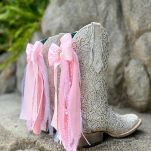 May include: A pair of white western-style boots featuring an intricate, raised scrollwork pattern across the shaft. Each boot is adorned with a large, light pink satin bow attached to the side, featuring cascading ribbons, lace trim, and small white beads.