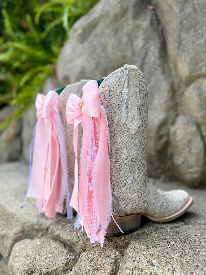 May include: A pair of white western-style boots featuring an intricate, raised scrollwork pattern across the shaft. Each boot is adorned with a large, light pink satin bow attached to the side, featuring cascading ribbons, lace trim, and small white beads.