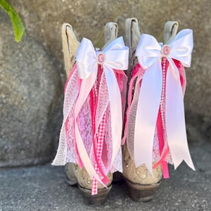 May include: A pair of beige cowboy boots adorned with white satin bows, pink ribbons, and lace. Each bow features a pink jewel. The boots have decorative stitching and are set against a neutral background.