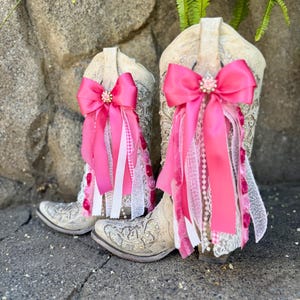 Op de afbeelding: Een paar off-white cowboylaarzen met zilveren borduursels. Elke laars is versierd met een grote, felroze strik met een centraal juweel. Roze en witte linten en kant hangen naar voren, wat een decoratief effect geeft.