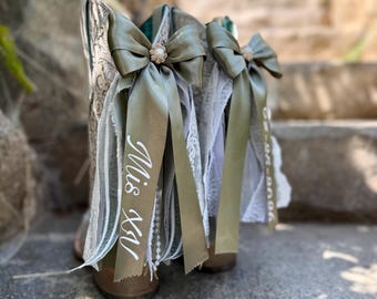 Peut inclure: Une paire de bottes de cow-boy marron clair ornées de nœuds en satin vert olive avec des perles centrales. Les rubans portent l'inscription blanche Mis XV et une date, agrémentés de dentelle blanche et de rangées de perles.