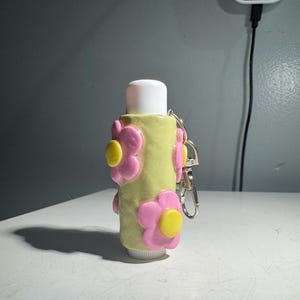 May include: A light green lip balm tube decorated with pink and yellow flower accents. The tube has a white cap and a silver keychain attachment. The product is on a white surface.