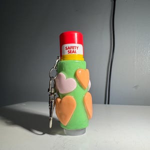 Könnte beinhalten: Ein grüner Lippenbalsam-Behälter mit rosa und orangefarbenen Herzen, gekrönt von einer roten Kappe mit der Aufschrift "SAFETY SEAL". Er hat einen silbernen Schlüsselanhänger. Der Behälter steht auf einer weißen Oberfläche.