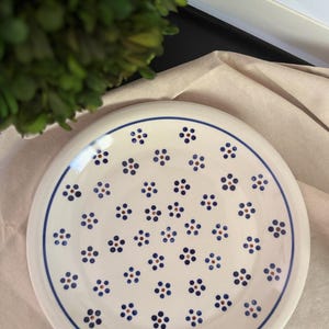 May include: A round ceramic plate with a cream-colored base featuring a hand-painted pattern of small blue flowers with brown centers. A single thin blue line circles the rim of the plate, framing the repeating floral motif across the surface.