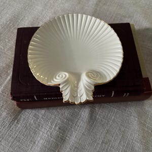 May include: A cream-colored ceramic dish shaped like a scallop shell with a gold-trimmed edge. The dish features a ribbed, fan-like texture radiating from the center toward the scalloped rim, with two decorative spiral accents at the base.