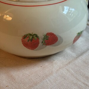Peut inclure: Une th&eacute;i&egrave;re en c&eacute;ramique de couleur cr&egrave;me orn&eacute;e d'un motif de fraises rouges avec des feuilles vertes. Le couvercle et le corps de la th&eacute;i&egrave;re sont soulign&eacute;s par une fine ligne rouge horizontale. La surface de la c&eacute;ramique est lisse et brillante.