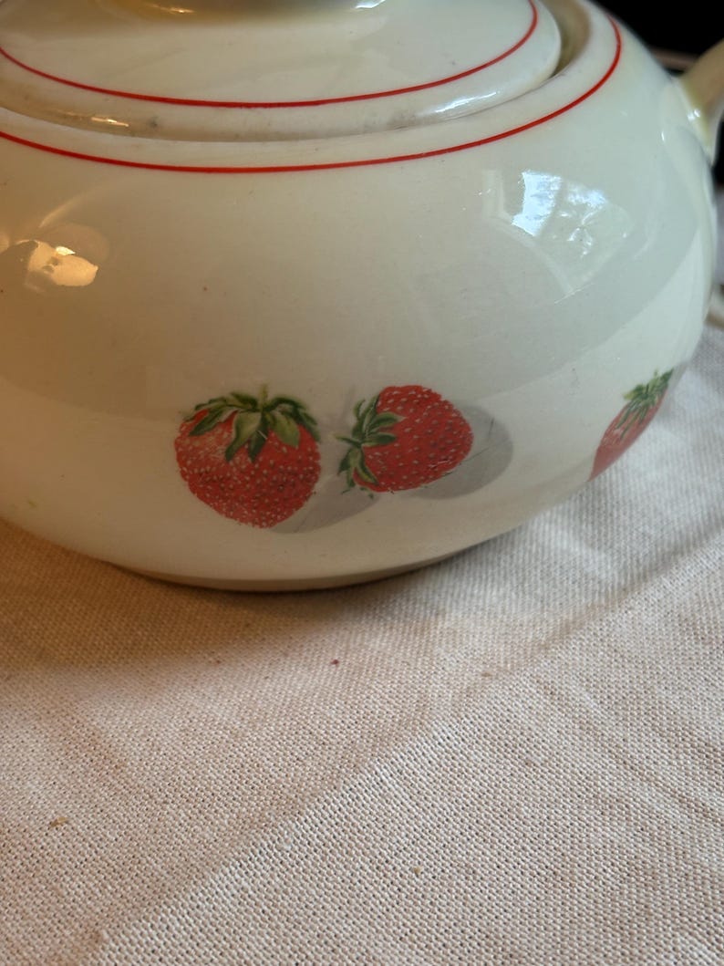 Peut inclure: Une th&eacute;i&egrave;re en c&eacute;ramique de couleur cr&egrave;me orn&eacute;e d'un motif de fraises rouges avec des feuilles vertes. Le couvercle et le corps de la th&eacute;i&egrave;re sont soulign&eacute;s par une fine ligne rouge horizontale. La surface de la c&eacute;ramique est lisse et brillante.