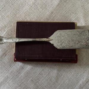 May include: A silver-toned metal cake server featuring an ornate, embossed floral pattern on the flat blade and a decorative shell-like motif on the handle. The utensil has a long, slender neck connecting the handle to the wide, rectangular blade.
