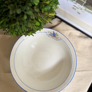 May include: A round white ceramic bowl featuring a thin blue rim and a small decorative illustration of a blue bird perched on a branch near the edge. The bowl has a smooth, glossy finish and a simple, clean design suitable for serving food.