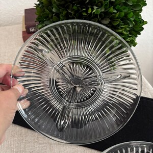May include: Clear glass serving dish with a divided design. The dish has a radial pattern emanating from the center, creating multiple sections. The dish is round and appears to be vintage. A hand is holding the edge of the dish.