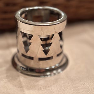 May include: A cylindrical silver-toned metal candle holder featuring a cut-out design of evergreen trees around the perimeter. The object has a smooth, reflective finish with a solid circular base and a matching rim at the top edge.