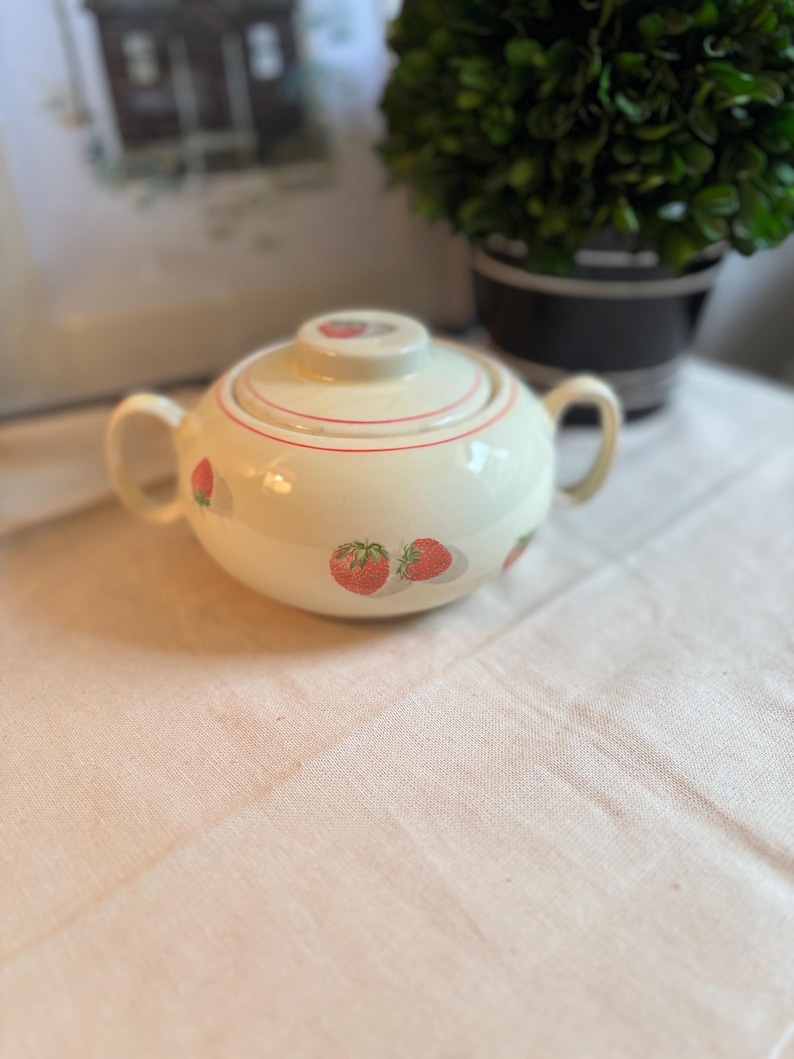 May include: A cream-colored ceramic sugar bowl with two side handles and a matching lid. The bowl features a repeating pattern of red strawberries with green leaves and thin red circular accent lines around the lid and the body of the bowl.