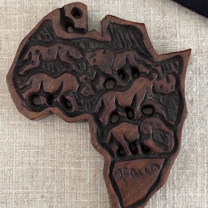 May include: A carved wooden pendant in the shape of the African continent. The dark brown pendant features carved images of animals and the word "Africa". The pendant has several holes.
