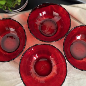 May include: A set of four deep red glass bowls with a scalloped rim design. Each bowl features a translucent finish and a circular base. The word Arcoroc is embossed in the center of each bowl, indicating the brand of this glassware set.