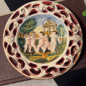 May include: A decorative ceramic plate with a detailed relief design. The plate features a central scene of cherubic figures in a garden setting, surrounded by a red and white openwork border. The rim is trimmed with gold accents.