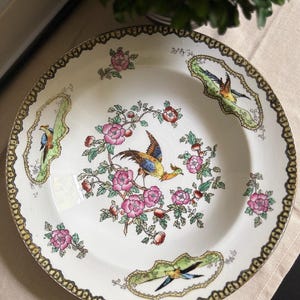 May include: A round ceramic plate with a cream-colored base featuring a central illustration of a colorful pheasant surrounded by pink flowers and green leaves. The rim is decorated with a dark patterned border and three inset vignettes of birds.