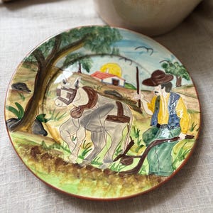 May include: A round ceramic plate featuring a hand-painted scene of a person in a yellow shirt and blue vest guiding a grey horse with a plow. The background depicts a rural landscape with a small house, a yellow sun, and green trees.
