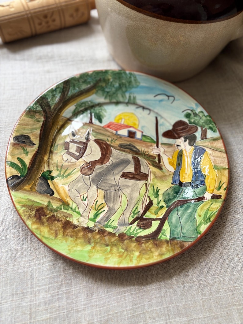 May include: A round ceramic plate featuring a hand-painted scene of a person in a yellow shirt and blue vest guiding a grey horse with a plow. The background depicts a rural landscape with a small house, a yellow sun, and green trees.
