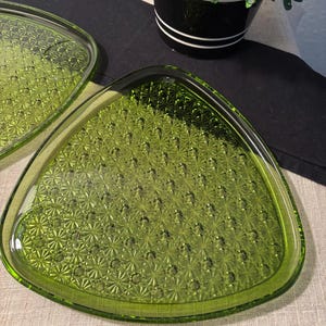 May include: Two green glass triangular serving dishes with a textured starburst pattern. The dishes are a vibrant green color and have a slightly raised edge. The dishes are likely vintage and suitable for serving appetizers or desserts.