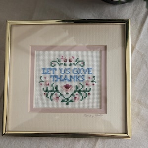 May include: A square cross-stitch embroidery in a gold-toned metal frame featuring the text LET US GIVE THANKS in blue, surrounded by a floral pattern in pink and green on a white fabric background with a light pink matting and a signature.