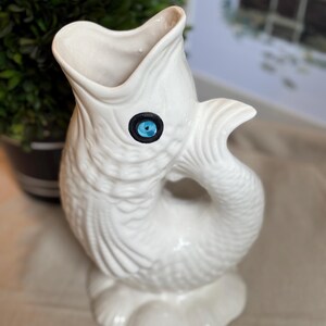 May include: A white ceramic pitcher shaped like a fish with an open mouth, textured scales, and a single bright blue eye with a black outline. The fish-shaped vessel features a curved tail handle and stands on a scalloped, shell-like base.