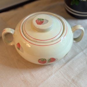 Peut inclure: Un sucrier en c&eacute;ramique de couleur cr&egrave;me avec deux anses et un couvercle assorti. La pi&egrave;ce pr&eacute;sente un motif r&eacute;p&eacute;titif de fraises rouges avec des feuilles vertes et de fines lignes circulaires rouges autour du couvercle et du corps.
