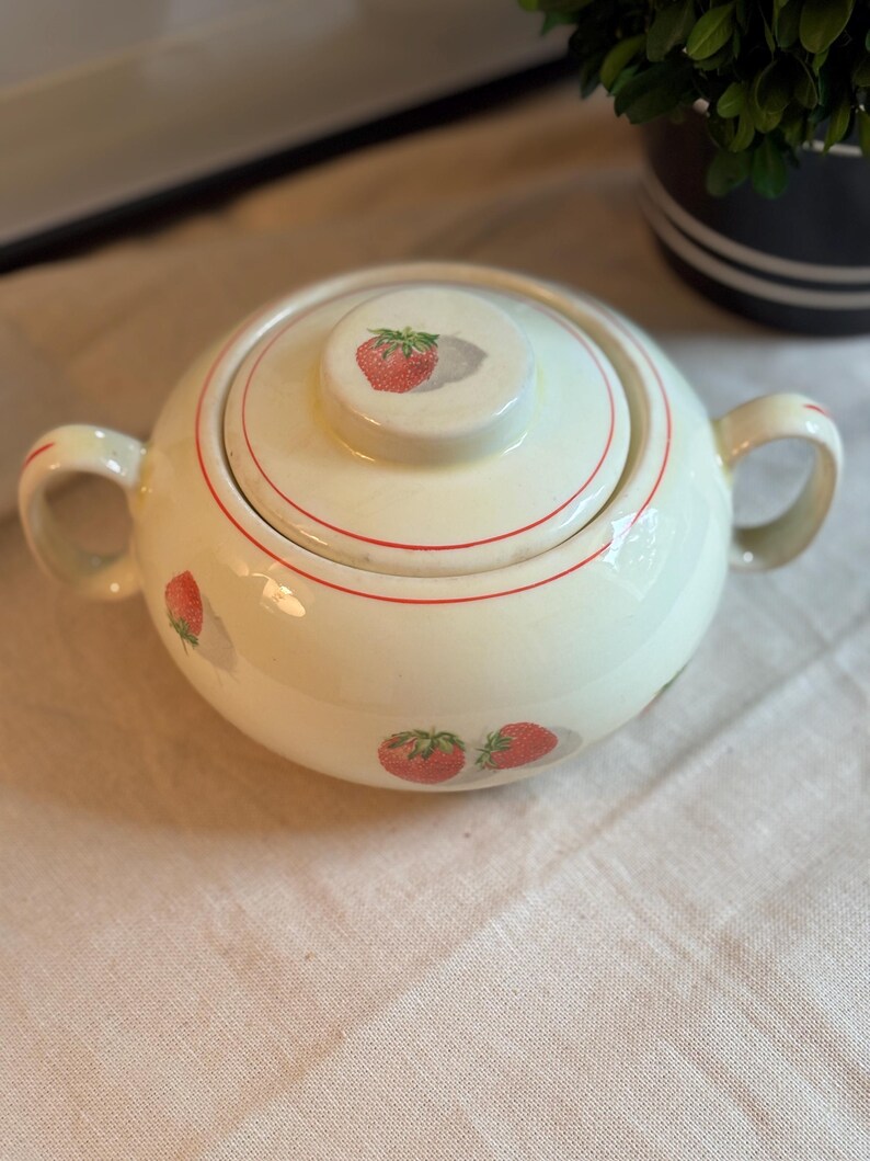 Peut inclure: Un sucrier en c&eacute;ramique de couleur cr&egrave;me avec deux anses et un couvercle assorti. La pi&egrave;ce pr&eacute;sente un motif r&eacute;p&eacute;titif de fraises rouges avec des feuilles vertes et de fines lignes circulaires rouges autour du couvercle et du corps.