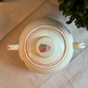 Peut inclure: Un sucrier en c&eacute;ramique de couleur cr&egrave;me avec un couvercle assorti, orn&eacute; d'une illustration de fraise rouge au centre du couvercle et de fines lignes circulaires rouges. Le bol poss&egrave;de deux anses lat&eacute;rales, chacune d&eacute;cor&eacute;e d'un trait rouge.