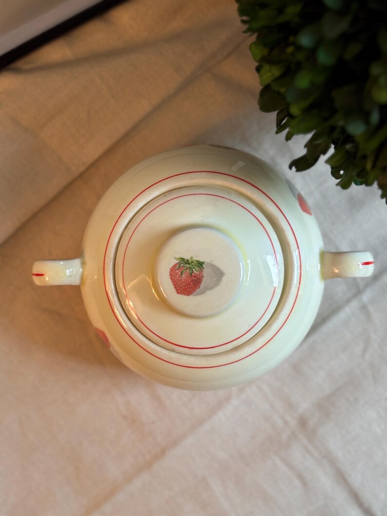 Peut inclure: Un sucrier en c&eacute;ramique de couleur cr&egrave;me avec un couvercle assorti, orn&eacute; d'une illustration de fraise rouge au centre du couvercle et de fines lignes circulaires rouges. Le bol poss&egrave;de deux anses lat&eacute;rales, chacune d&eacute;cor&eacute;e d'un trait rouge.