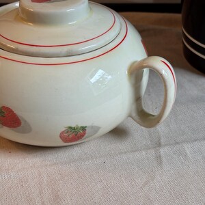Peut inclure: Un sucrier en c&eacute;ramique de couleur cr&egrave;me avec un couvercle assorti, orn&eacute; d'un motif r&eacute;p&eacute;titif de fraises rouges avec des feuilles vertes et de fines lignes circulaires rouges. Le bol comprend une anse incurv&eacute;e sur le c&ocirc;t&eacute; et un couvercle rond.