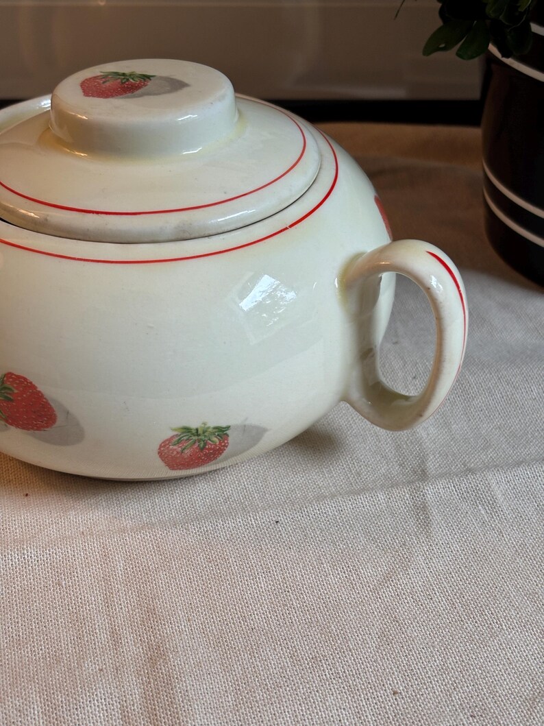 Peut inclure: Un sucrier en c&eacute;ramique de couleur cr&egrave;me avec un couvercle assorti, orn&eacute; d'un motif r&eacute;p&eacute;titif de fraises rouges avec des feuilles vertes et de fines lignes circulaires rouges. Le bol comprend une anse incurv&eacute;e sur le c&ocirc;t&eacute; et un couvercle rond.