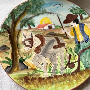 May include: A round ceramic plate featuring a hand-painted scene of a person in a brown hat, yellow shirt, and blue vest guiding a grey donkey through a field. The background includes a small white house with a red roof under a yellow sun.
