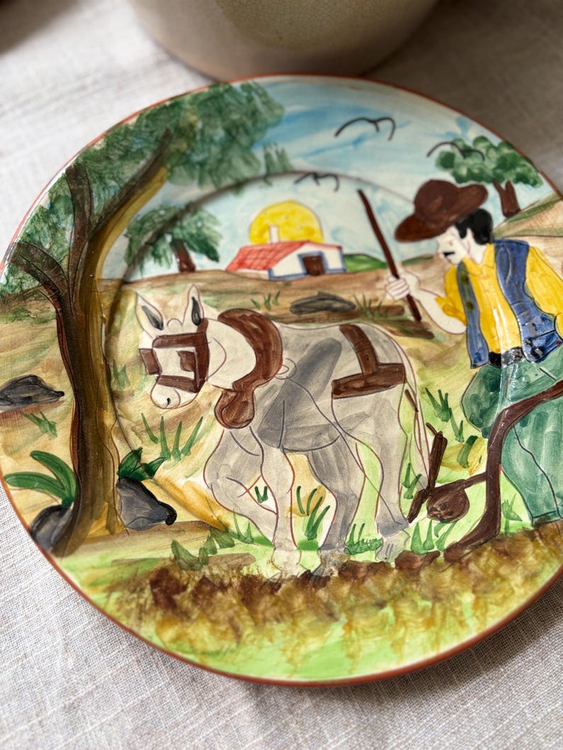 May include: A round ceramic plate featuring a hand-painted scene of a person in a brown hat, yellow shirt, and blue vest guiding a grey donkey through a field. The background includes a small white house with a red roof under a yellow sun.